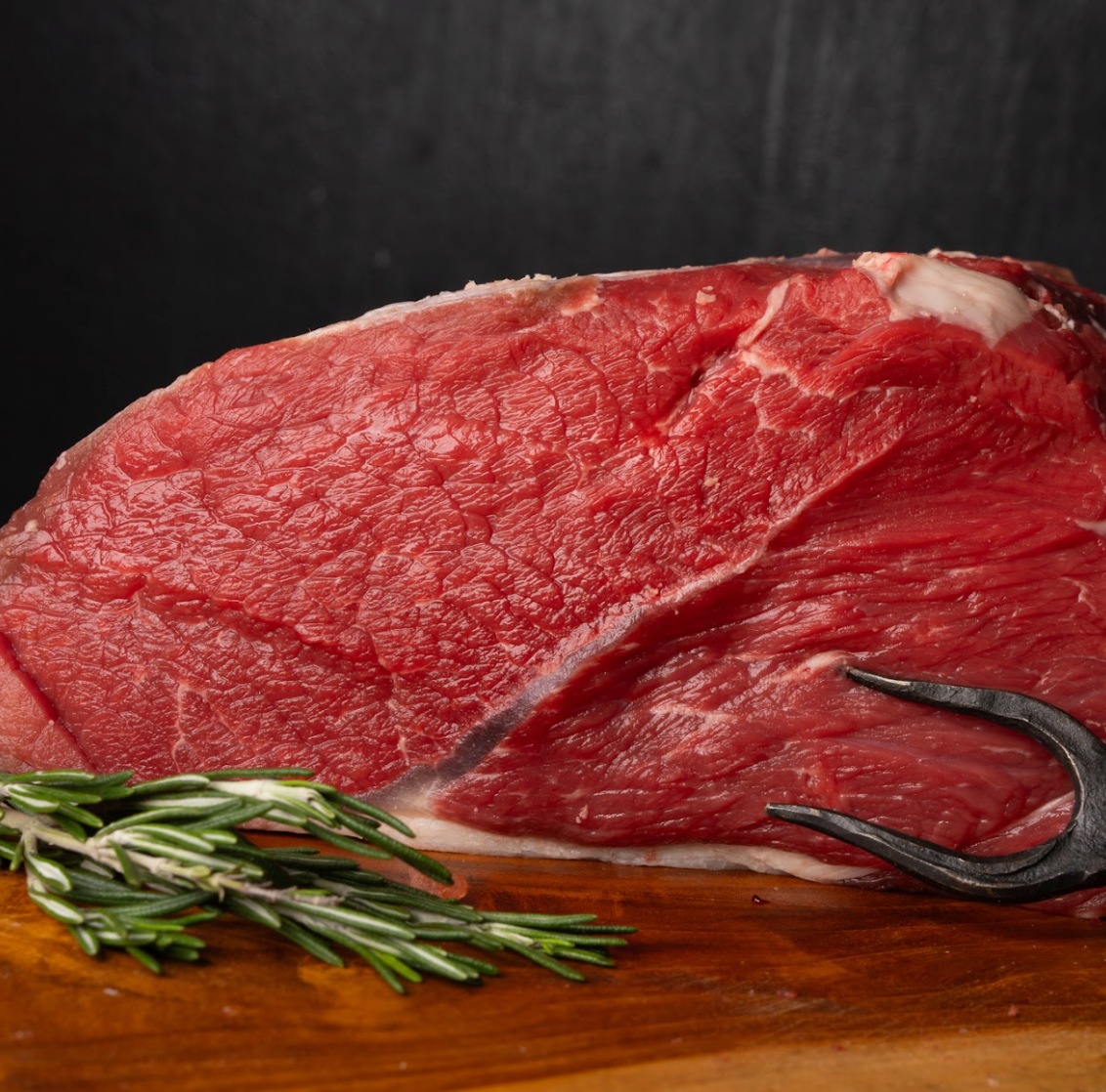 Raw steak on a wooden board with rosemary and a carving fork against a dark background