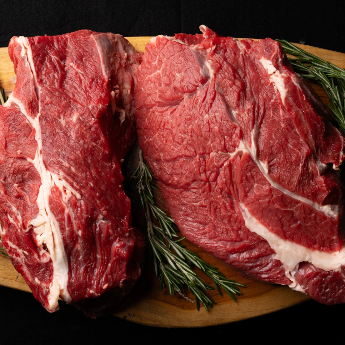 Two raw steaks on a wooden cutting board with rosemary sprigs against a dark background