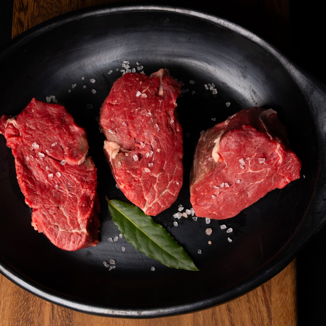 Three pieces of raw beef in a black pan with salt and a bay leaf on a wooden surface.