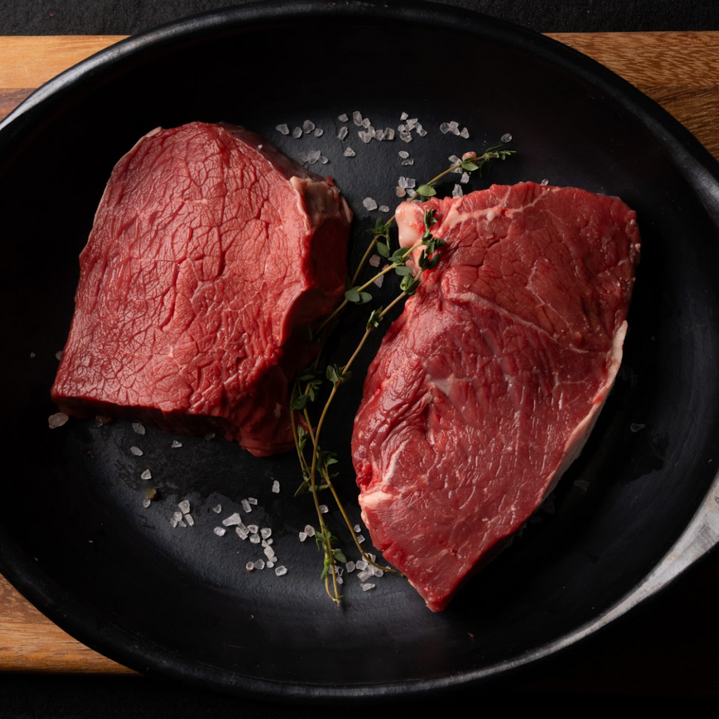 Two pieces of raw beef on a black plate with herbs and spices.