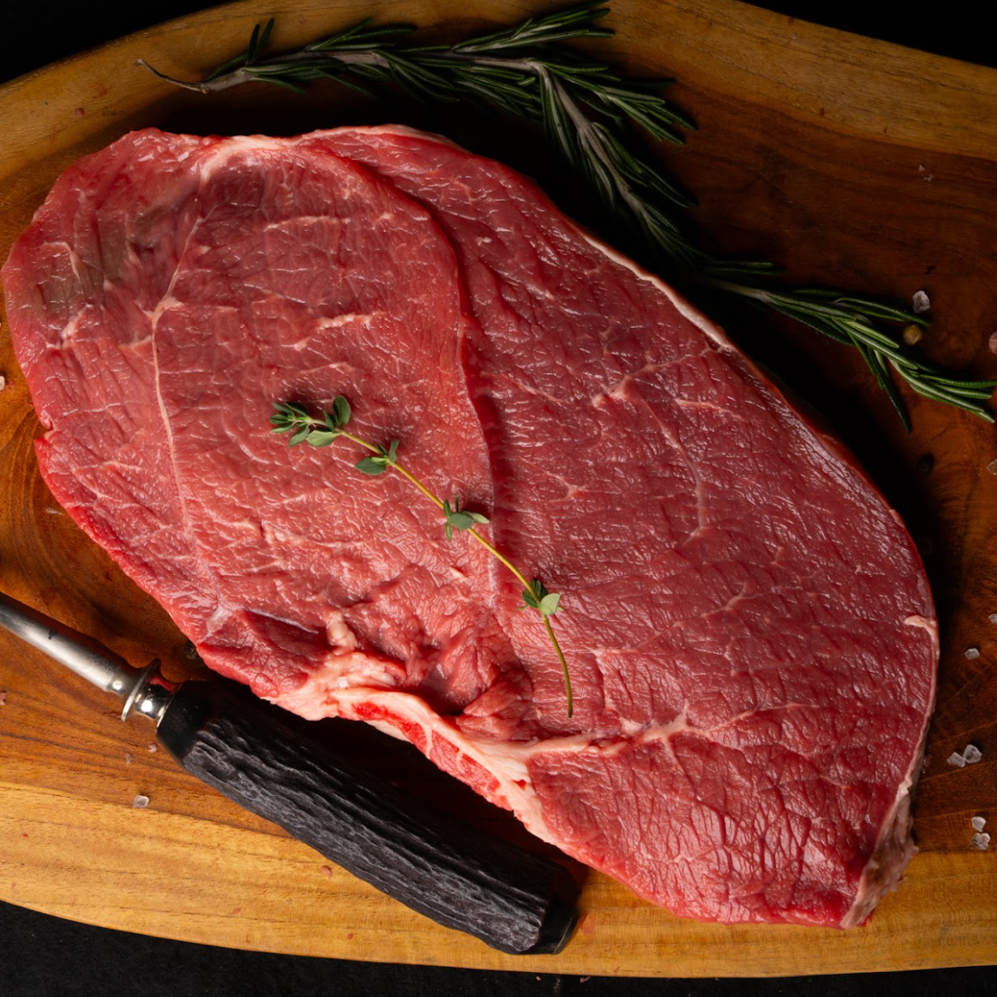 Raw steak on a wooden cutting board with herbs and a knife.